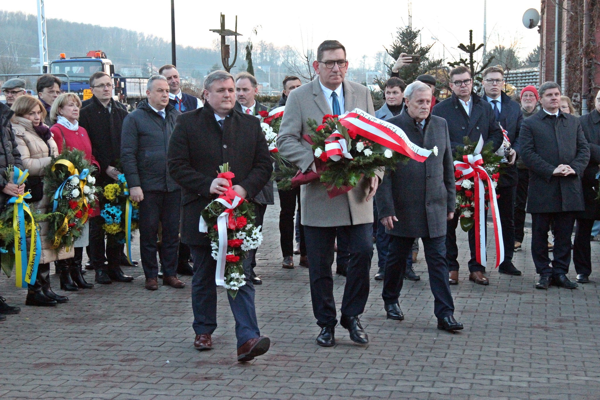 Trzech uczestników idzie złożyć wieńce przed tablice pamiątkową