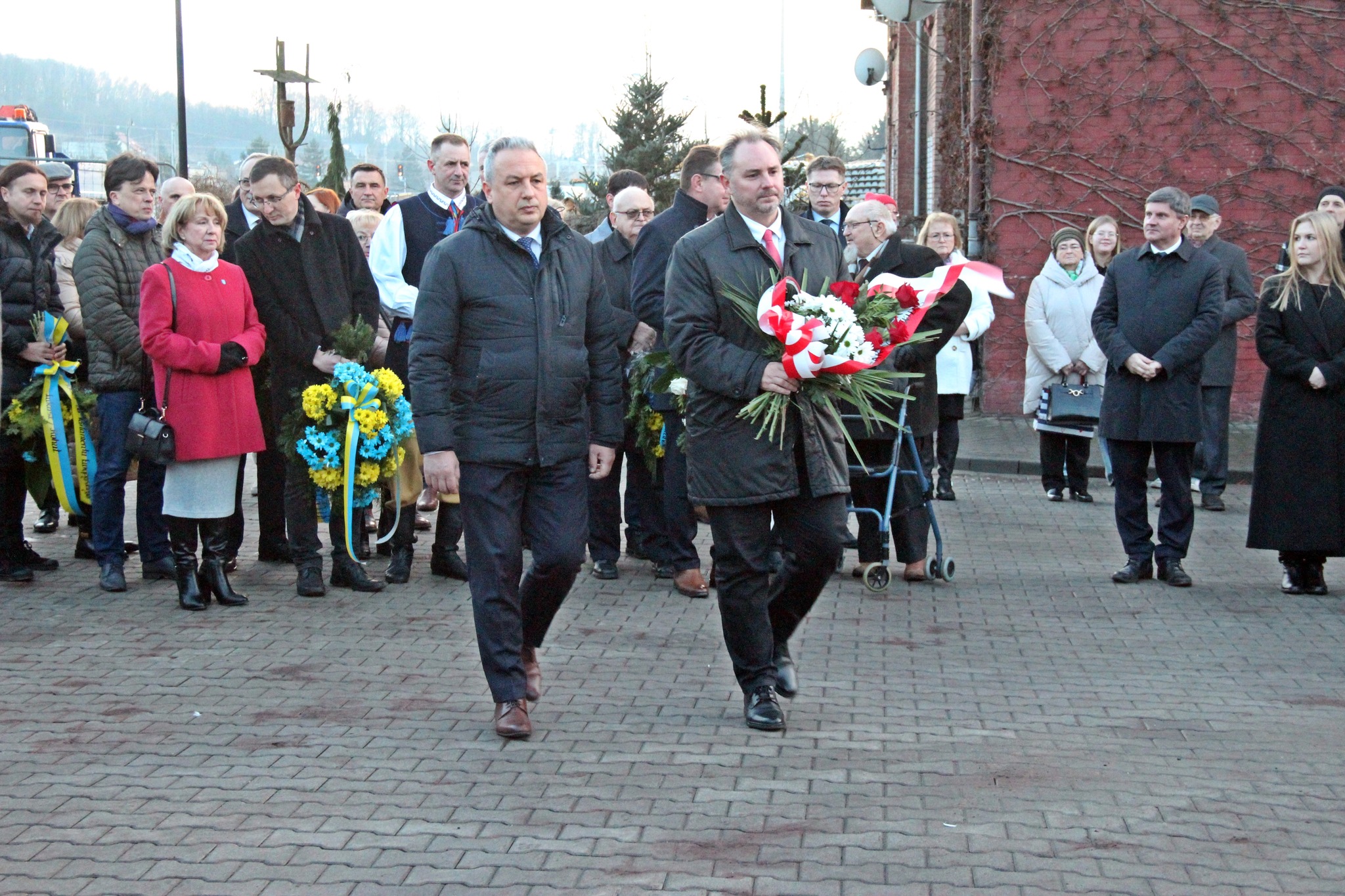 Dwóch uczestników wydarzenia idzie z wieńcem do tablicy pamiątkowej
