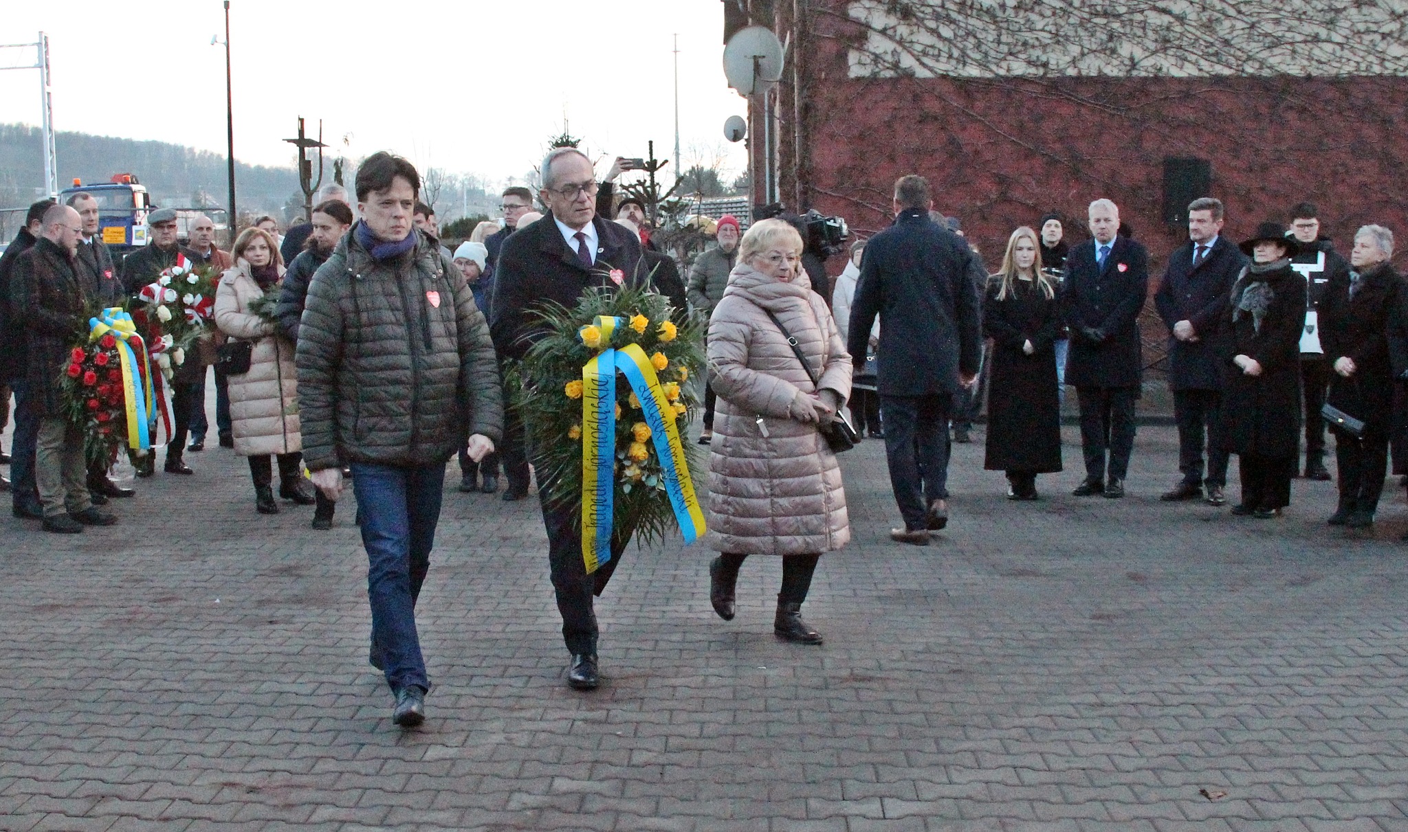 Trzy osoby idą z wieńcem do tablicy pamiątkowej