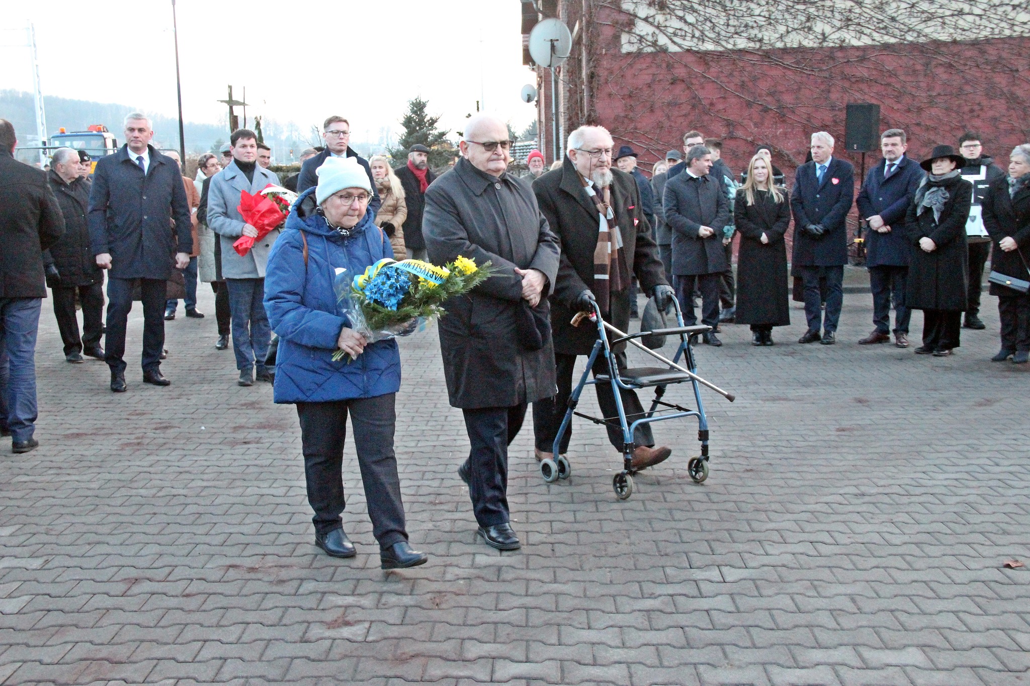 Trzy osoby podchodzą z wieńcem pod tablicę pamiątkową