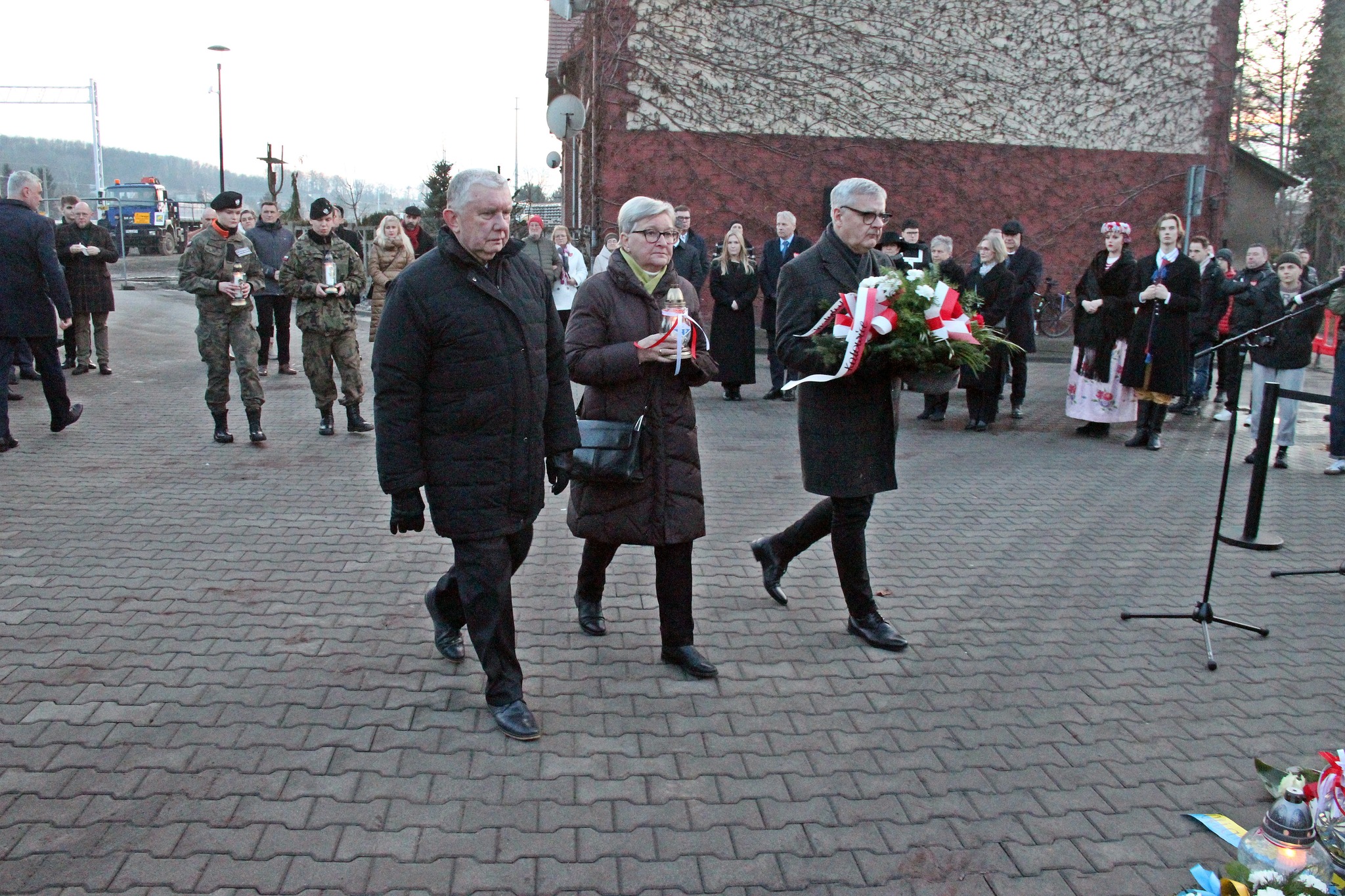 Trzy osoby podchodzą z wieńcem i zniczem pod tablicę pamiątkową